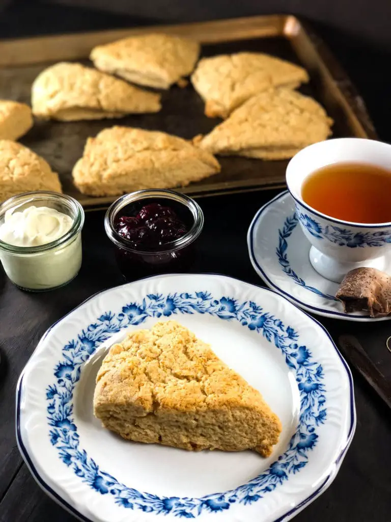 Scone made without butter in foreground, served over a white and blue place, a cup of tea besides and a baking sheet with fresh scones in the backgound